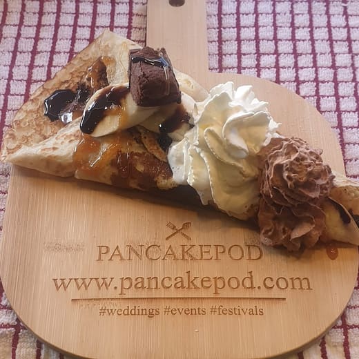 A Pancake Pod pancake folded on a wooden board with banana, chocolate sauce, caramel, a brownie piece, whipped cream and chocolate cream