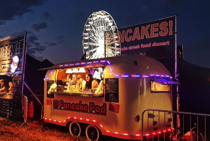 The Pancake Pod trailer glowing with festoon lights at night, serving customers beside a Ferris wheel